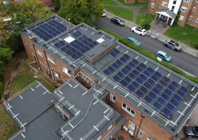 Aerial view of a brick building with extensive solar panels on its flat roof and surrounding street with cars and greenery nearby.