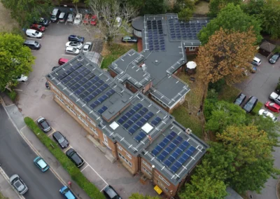 Aerial view of a brick building with extensive solar panels on the flat roofs, surrounded by parked cars and trees
