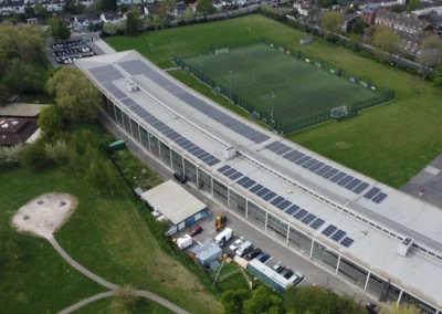 Aerial view of a solar-paneled building beside green soccer fields enclosed by a fence and a surrounding park and housing area.